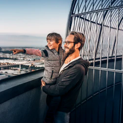 En man håller ett leende barn som pekar på stadsbilden från en hög utsiktsplattform med barer. Bakgrunden inkluderar höga byggnader och en flod.