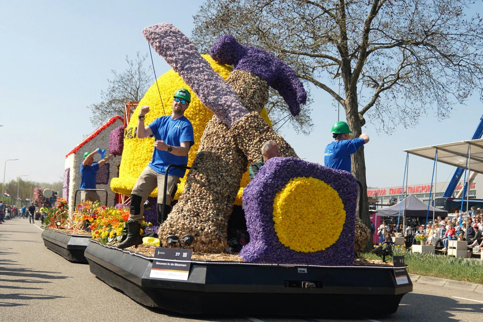 Οι άνδρες με μπλε πουκάμισα και πράσινα κράνη οδηγούν ένα άρμα διακοσμημένο με μεγάλα λουλούδια σε σχήμα σφυριού και θέμα κατασκευής.