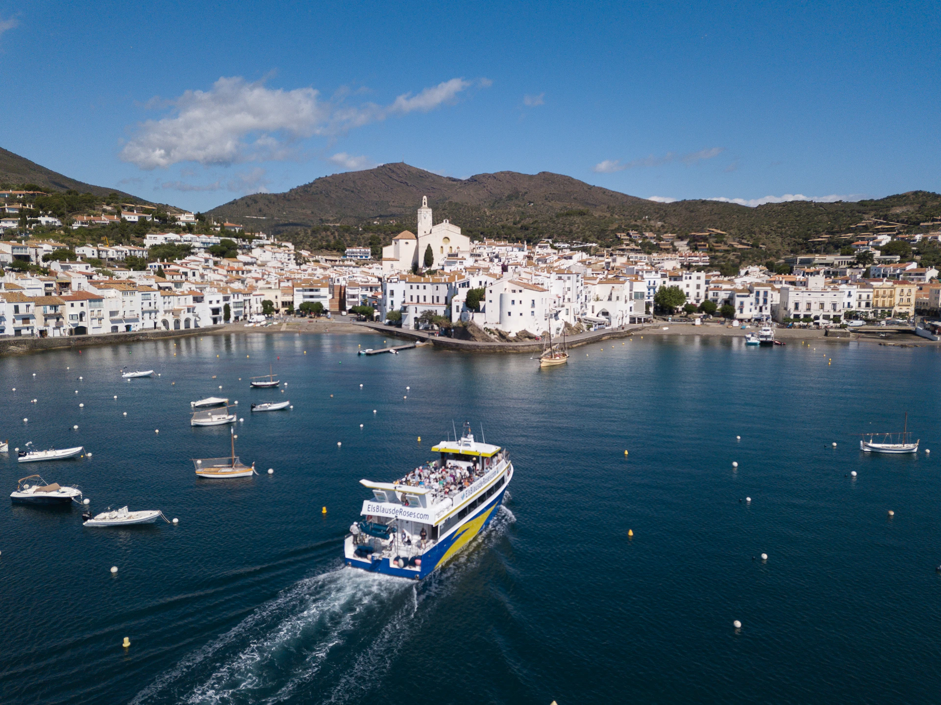 A boat sails on calm water in a coastal town with white buildings and a church against a backdrop of hills and blue sky.