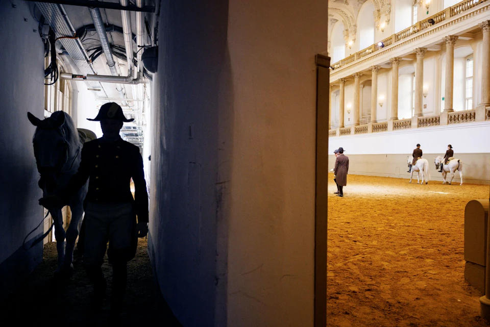 A person in a uniform and hat is in a dimly lit passage, while another person walks in a well-lit, ornate indoor equestrian arena.