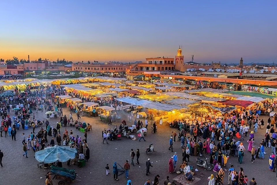 Evening market scene with numerous stalls, people walking and shopping, a lit tower, and buildings in the background under a dusk sky.