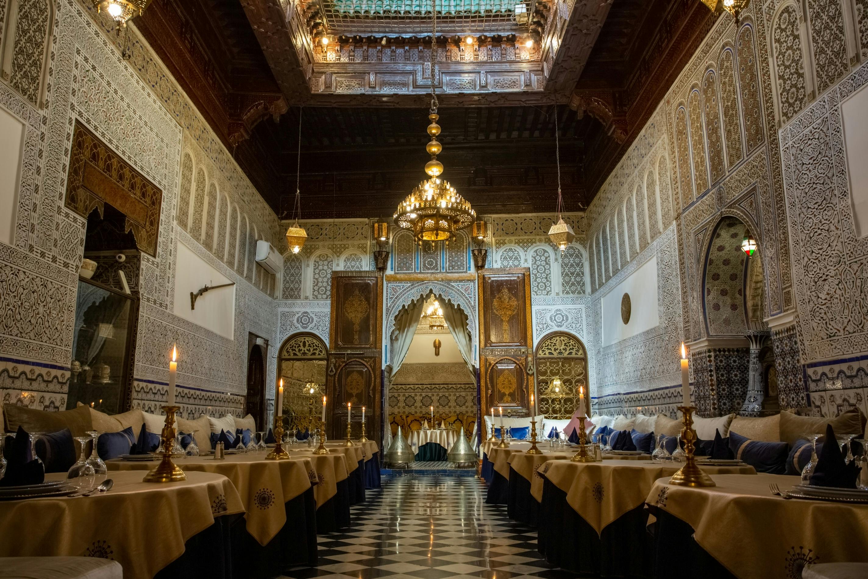Ornate restaurant interior with intricate Moroccan decor, chandeliers, patterned walls, ceiling, and rows of tables set for dining.