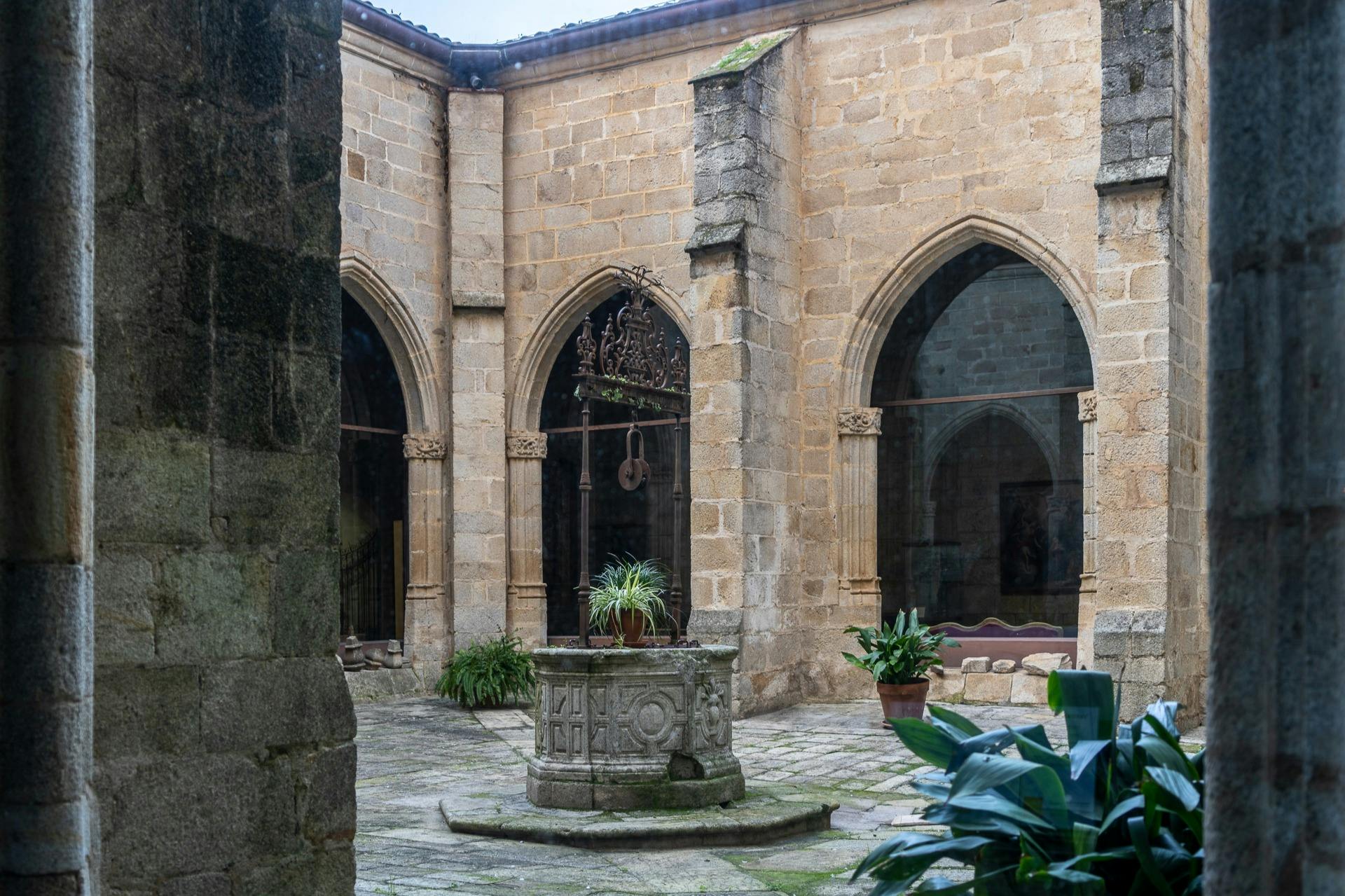 Een stenen binnenplaats met een decoratieve waterput als middelpunt, omringd door gotische bogen met potplanten en geplaveide vloeren.