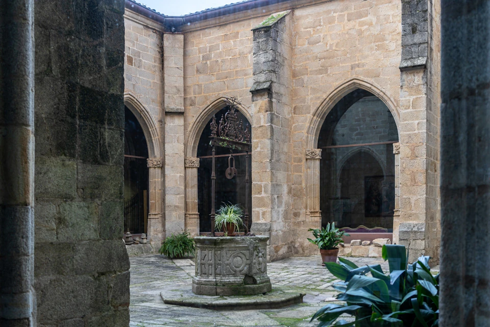 A stone courtyard with a central well, surrounded by arched windows and walls of a historic building. Plants are present.