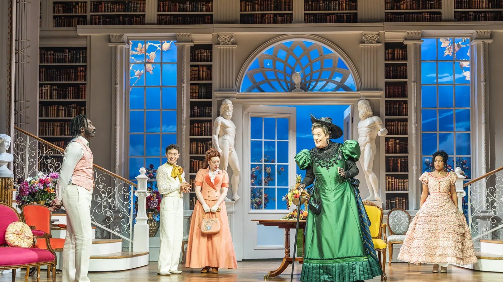 Τέσσερις ηθοποιοί με κοστούμια εποχής εκτελούν σε ένα περίτεχνο σκηνικό που μοιάζει με μια κομψή βιβλιοθήκη με αγάλματα και μια σκάλα.