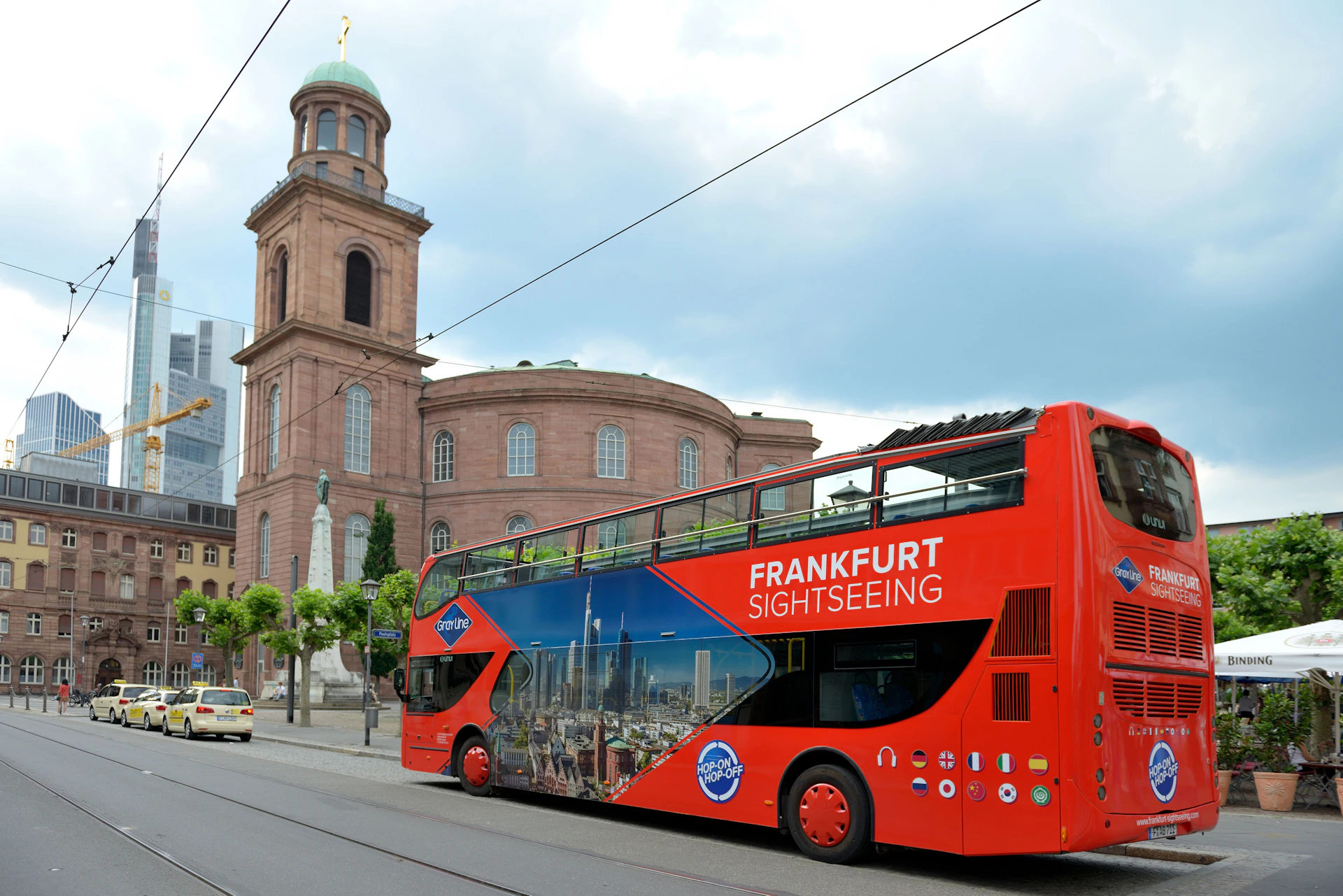 A red double-decker bus labeled "Frankfurt Sightseeing" near a historic brick building with a domed tower.