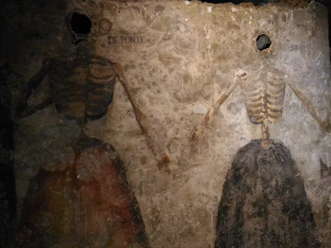 Two skeleton figures painted on a textured, aged wall, holding hands; partially visible text reads "di Ponti."