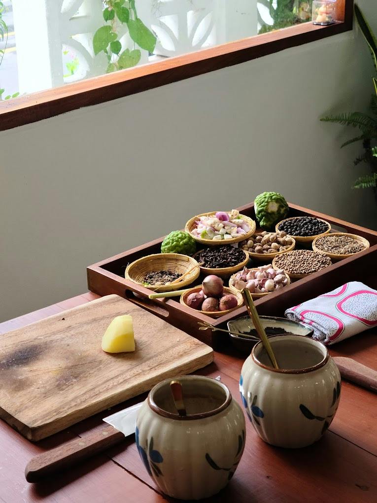 A wooden tray with various spices and ingredients next to jars and a chopping board with a piece of fruit.