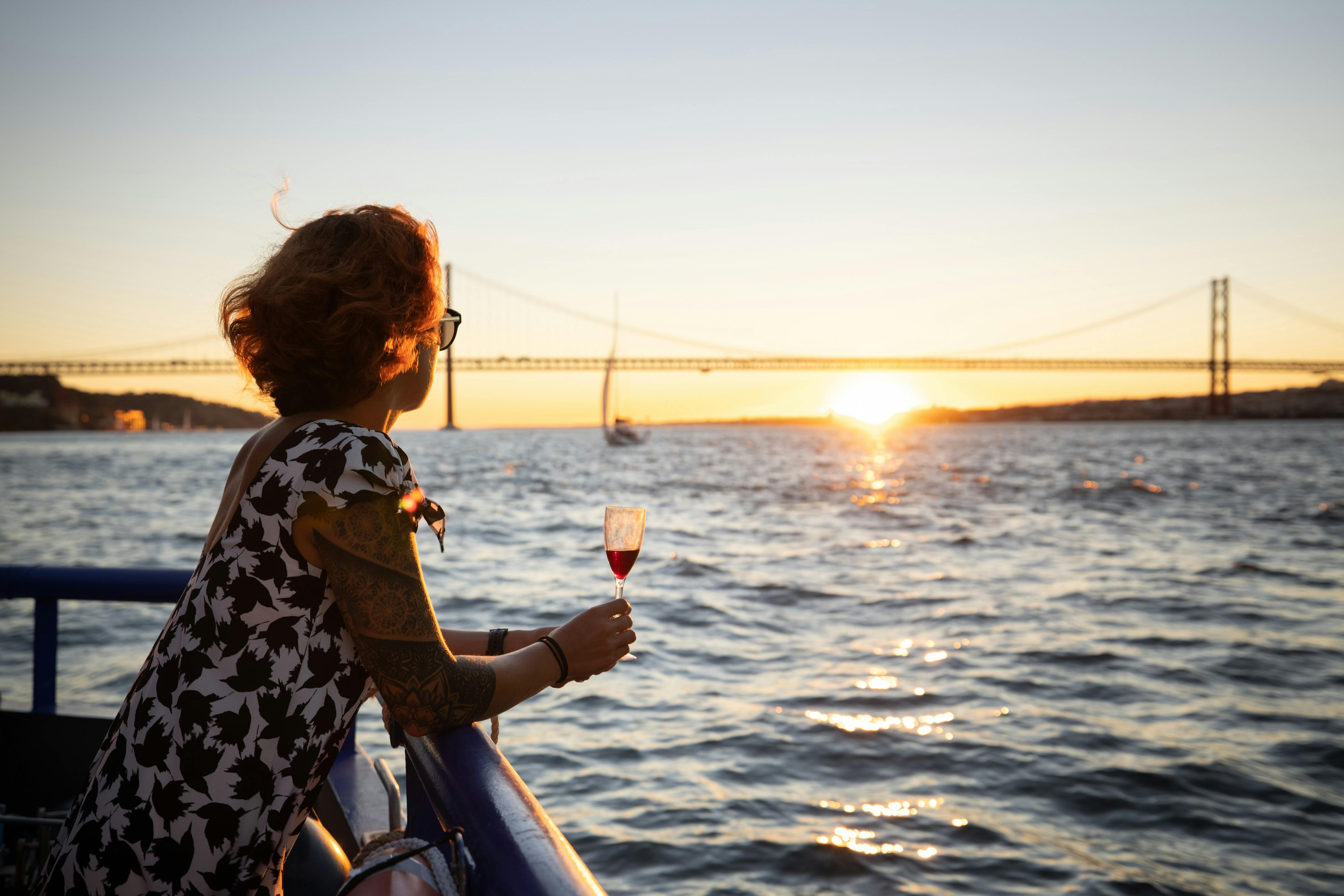 Persona con tatuaggi che tiene in mano un bicchiere di vino rosso, guardando un tramonto sull'acqua con un ponte sullo sfondo.