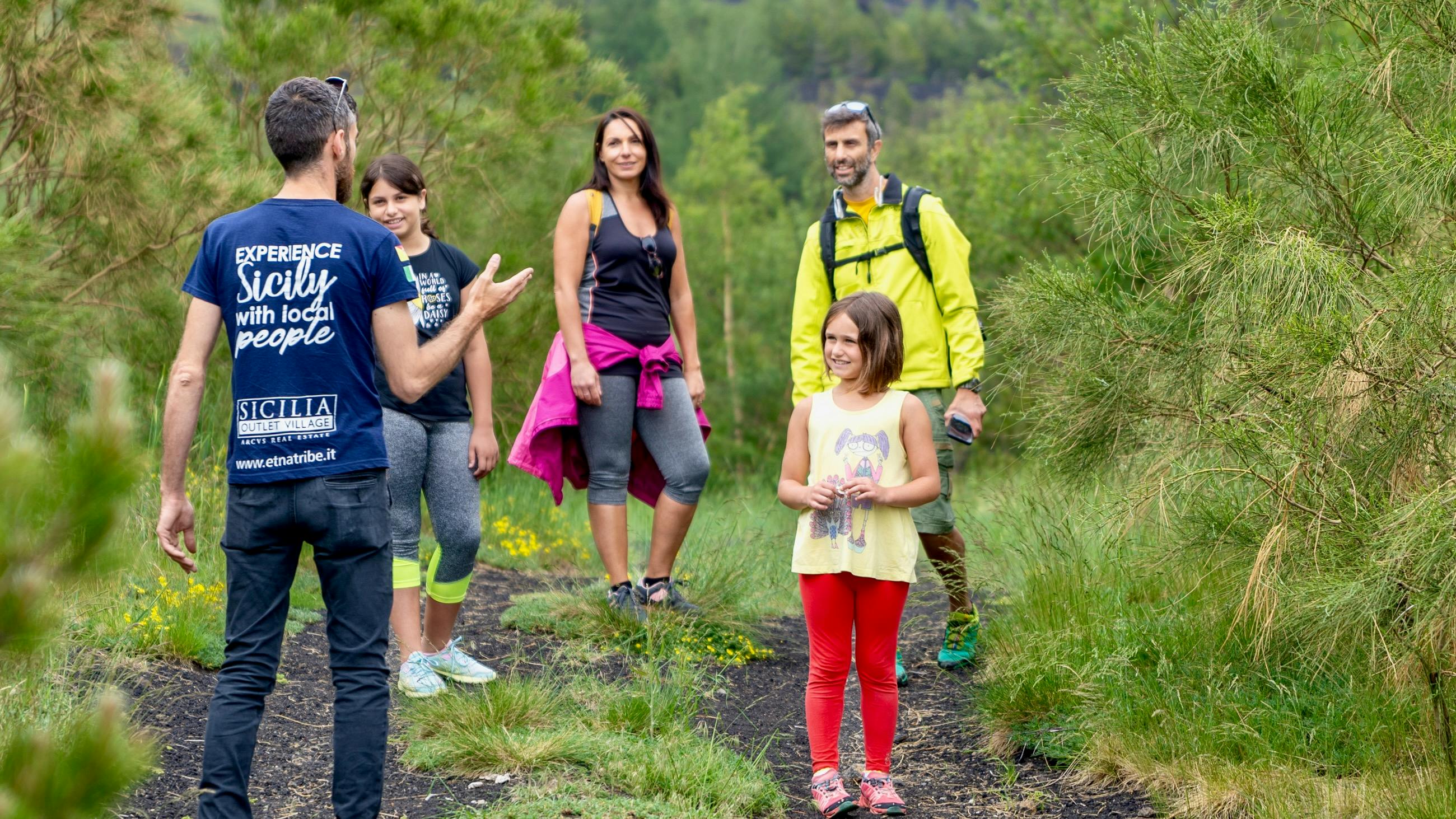 Een gids praat met een lachende groep van vier wandelaars, waaronder een kind, op een grassig pad met bomen op de achtergrond.