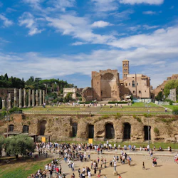 Folkmassor utforskar arkeologiska ruiner med kolumner och gamla strukturer under en delvis molnig himmel. Träd och grönska omger området.