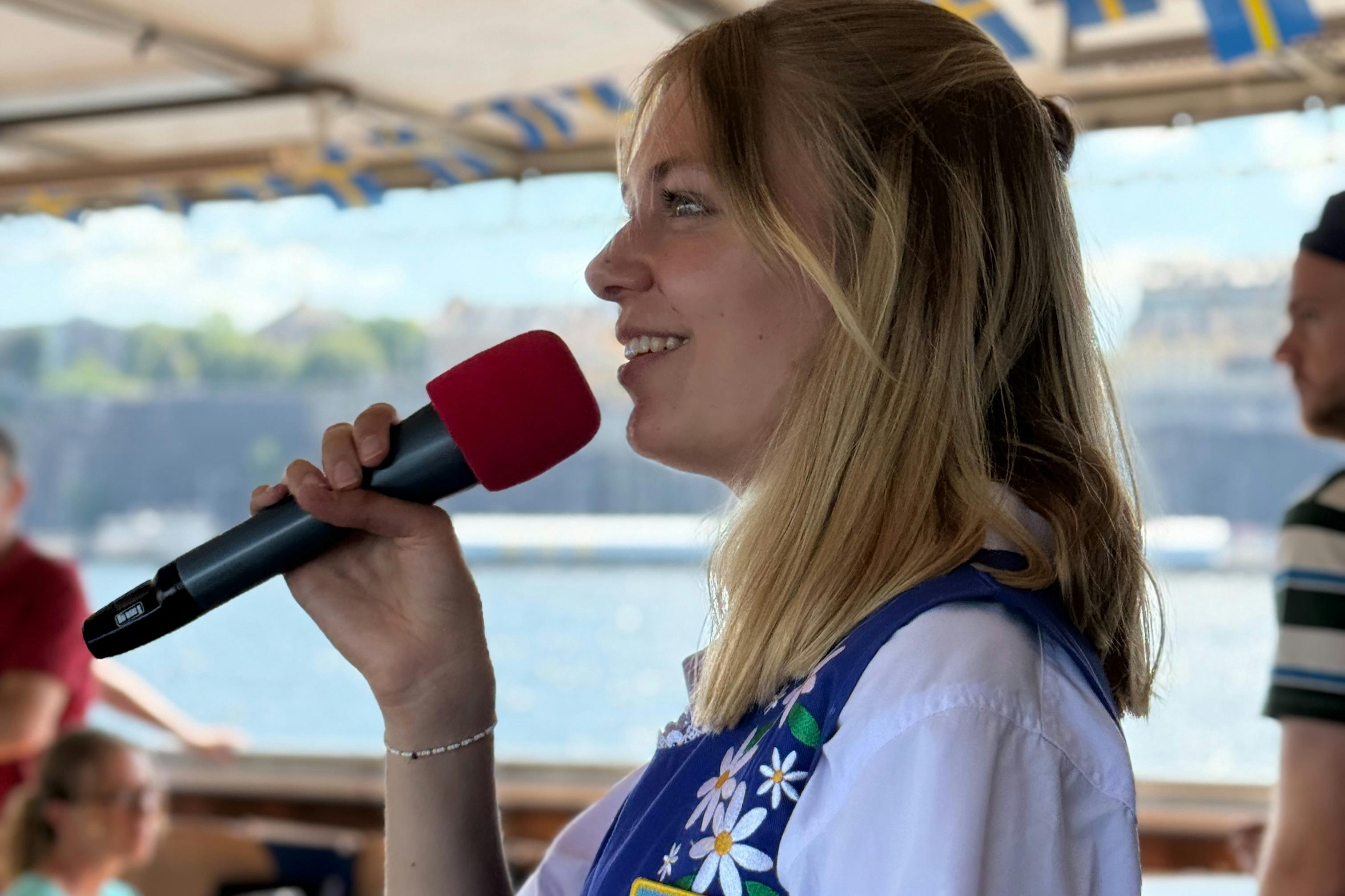 Donna che tiene un microfono con una copertura di schiuma rossa, sorride e parla su una barca con acqua e sfondo sfocato.