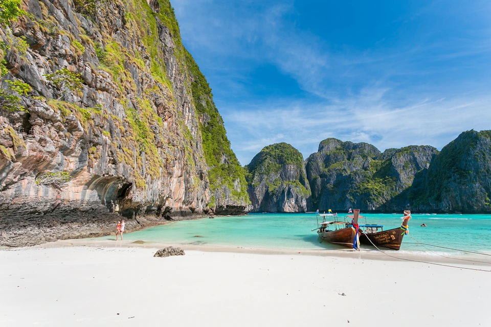 White sandy beach with turquoise water, a moored boat, steep rocky cliffs, lush greenery, and a clear blue sky.