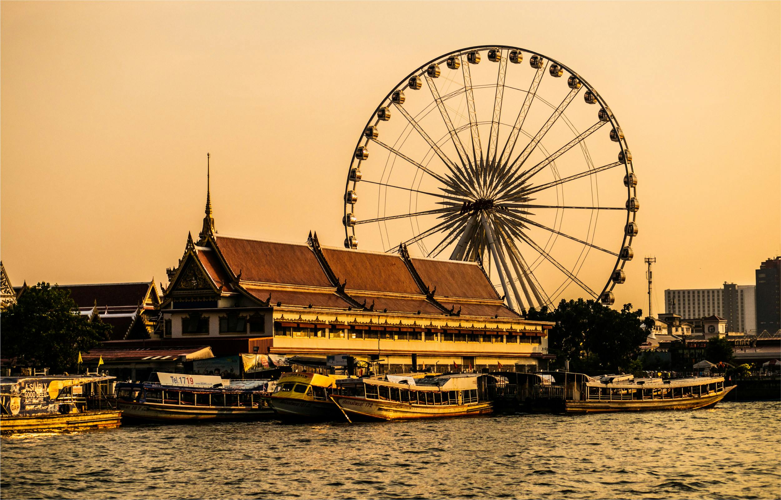 A riverside scene with a traditional building, boats docked in the foreground, and a ferris wheel in the background at sunset.