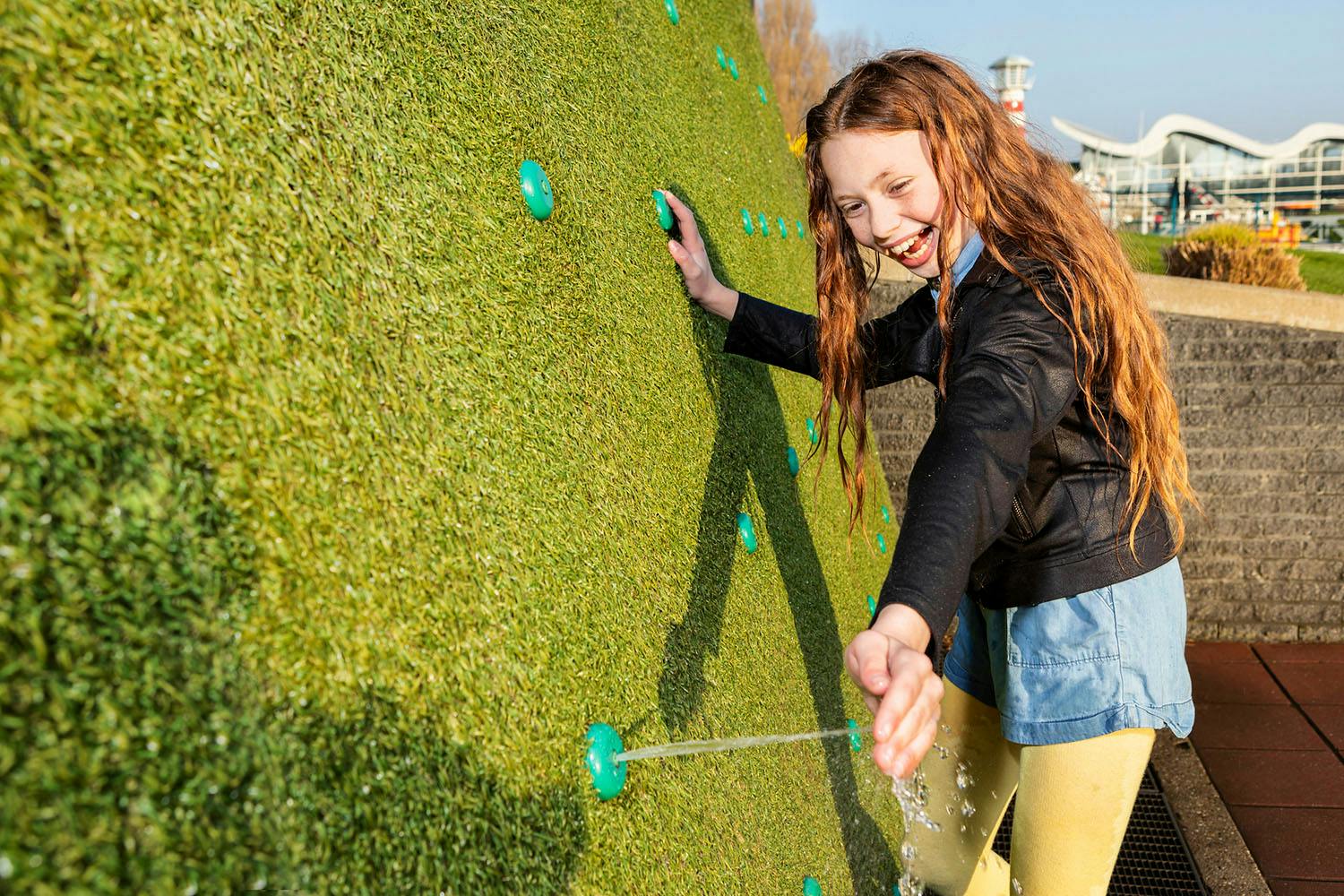 屋外の遊び場で青い噴水のある緑色の壁から水を飛ばしながら微笑んでいる少女。