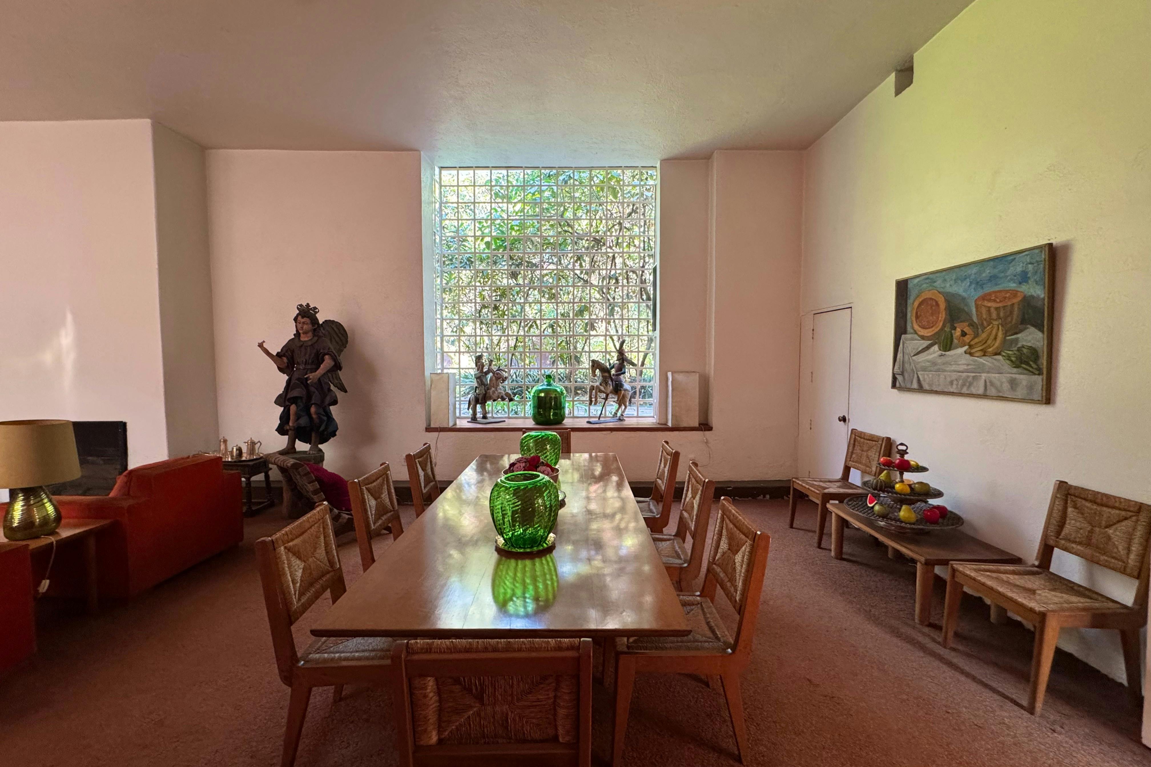 Una stanza con un grande tavolo da pranzo, vasi di vetro verde, sedie di vimini, una statua e un quadro di frutta alla parete.