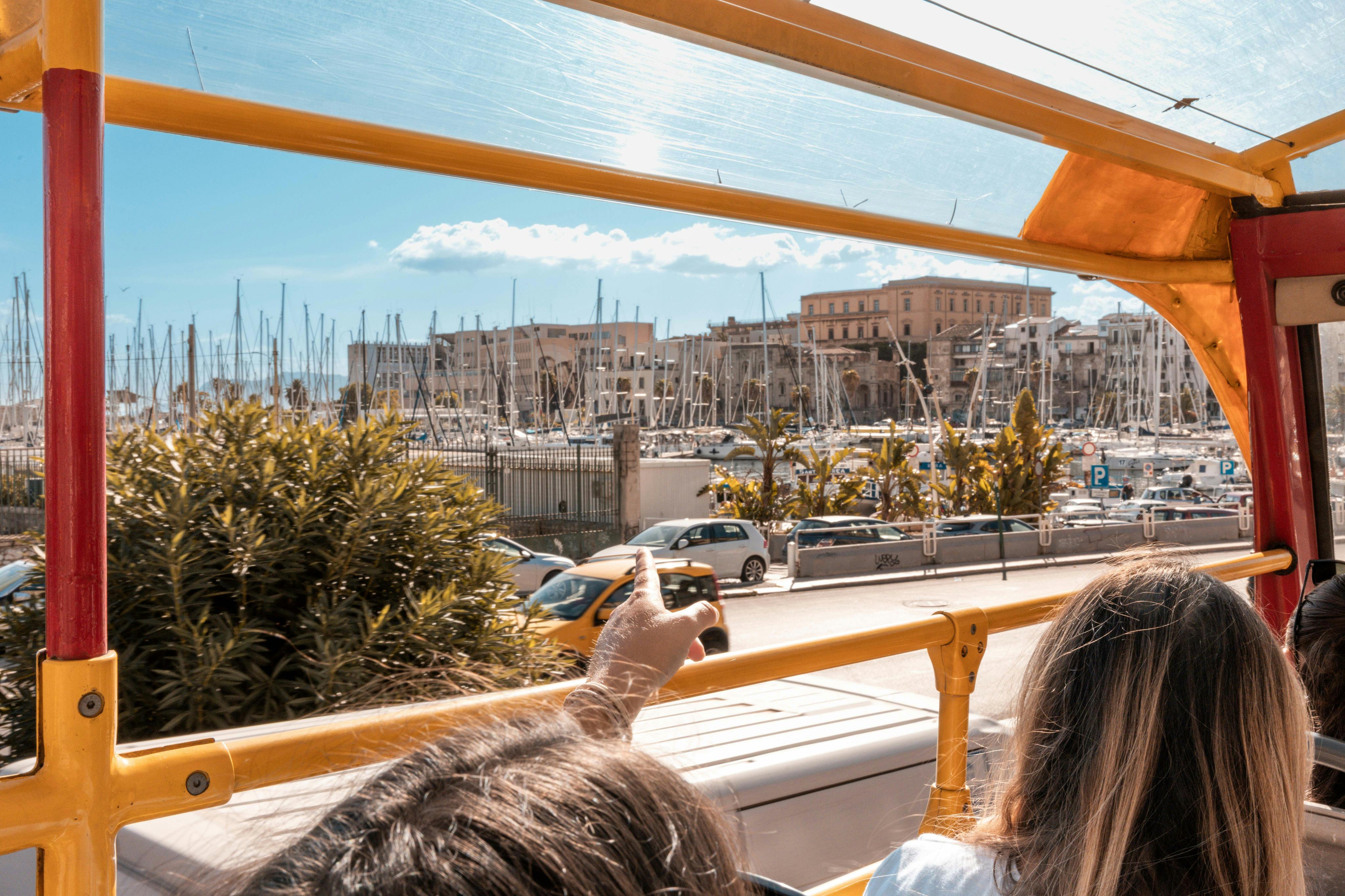 Persone sedute sul ponte superiore di un autobus turistico che indicano un porto con barche ed edifici sullo sfondo.