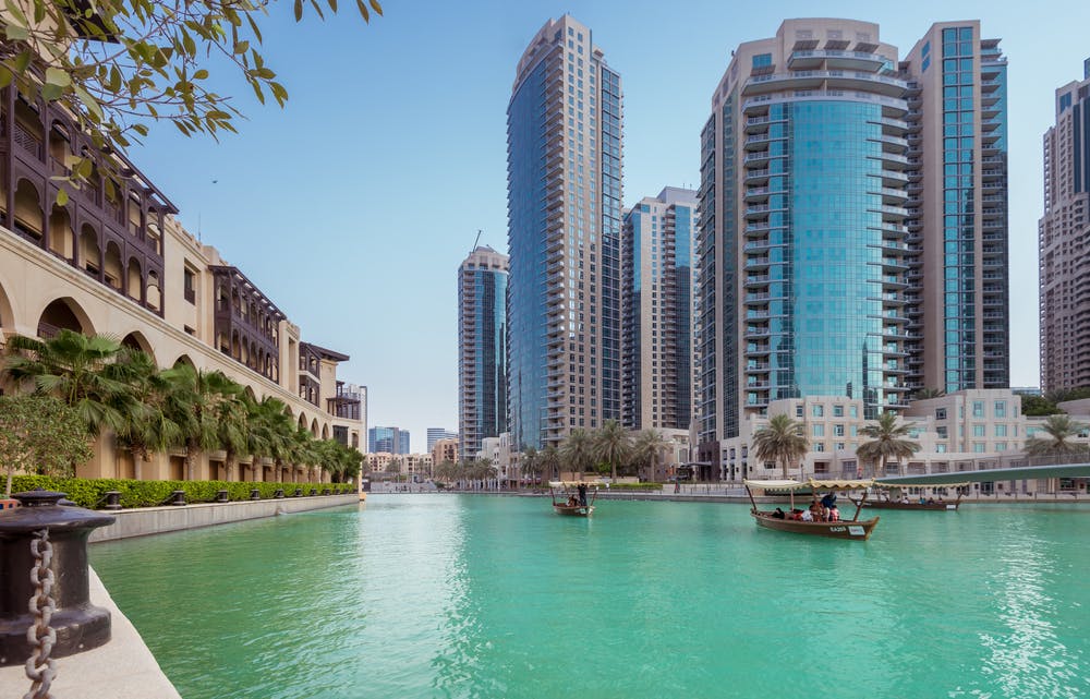 Canal de água azul-turquesa com pequenos barcos de madeira, ladeado por edifícios modernos altos e um edifício de estilo tradicional à esquerda. Céu azul claro.