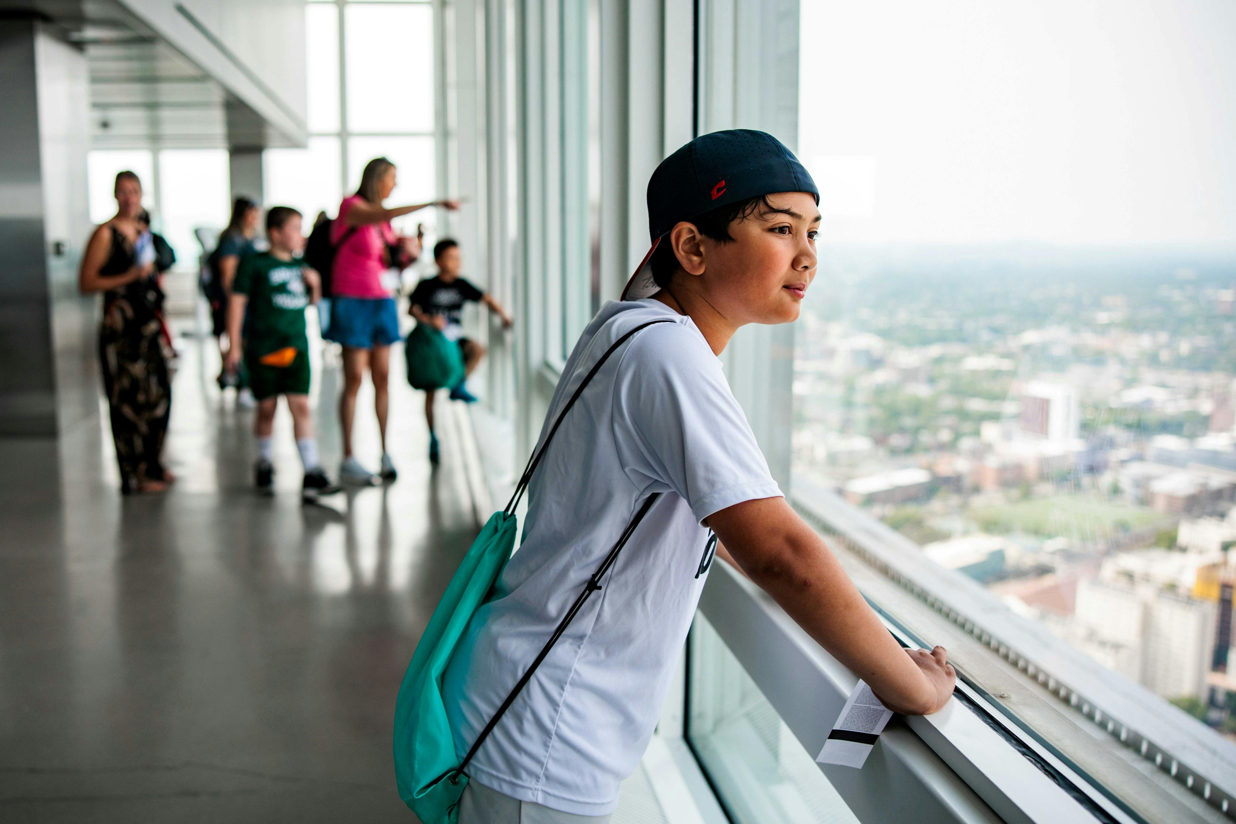 Un ragazzo con una camicia bianca e un cappellino da baseball guarda fuori da una grande vetrata di un grattacielo, con un paesaggio urbano sullo sfondo.