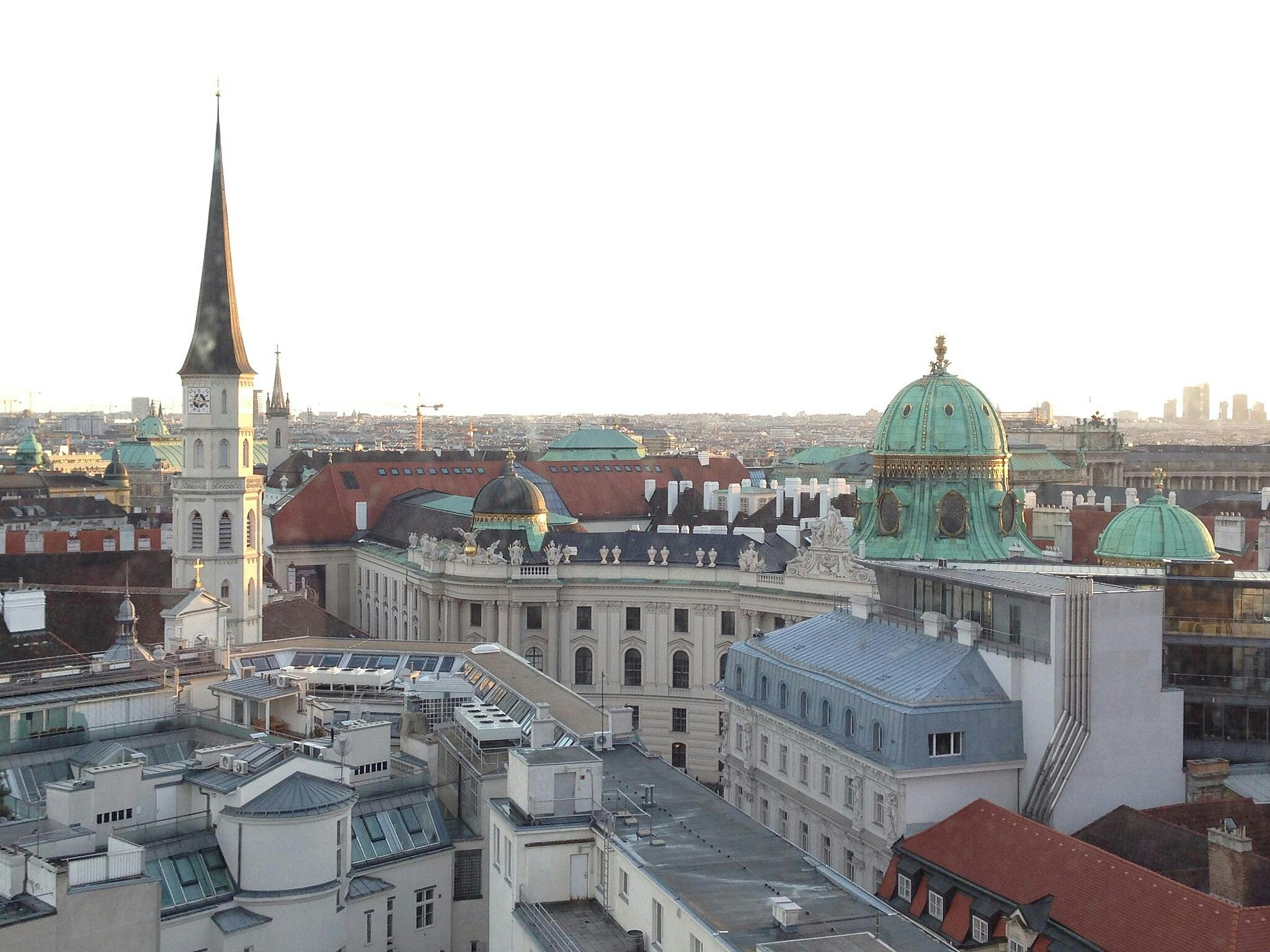 Panoráma města při západu slunce s historickými budovami s kopulemi a vysokou věží proti vzdálenému modernímu panoramatu.