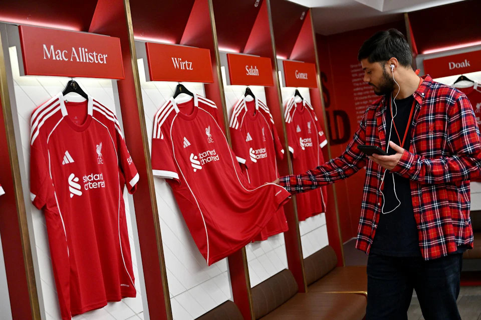 A man in a red plaid shirt inspects a red Liverpool FC jersey in a locker room with three other hanging jerseys labeled Mac Allister, Wirtz, Salah, and Gomez.