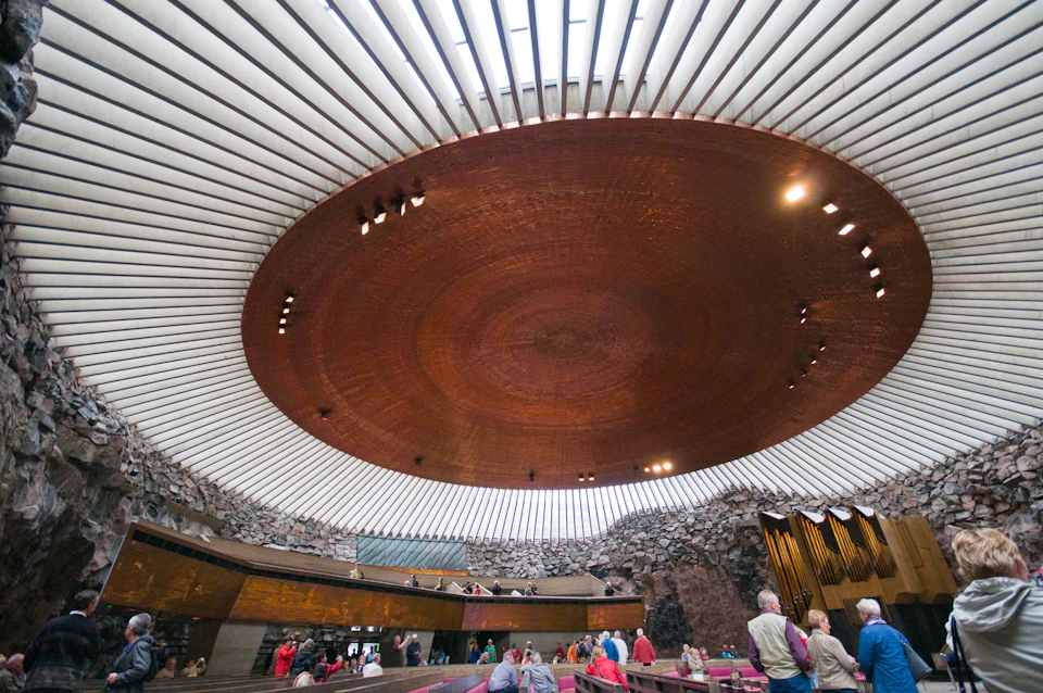 People inside a church with a large circular, copper ceiling and rugged stone walls. Elevated seating areas and organ pipes are visible.