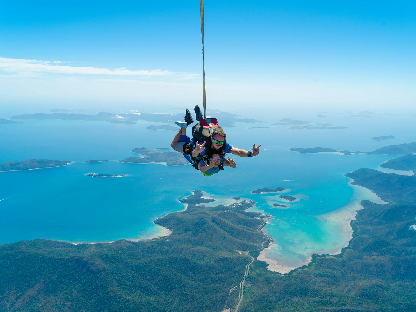 Dva lidé tandemově skáčou nad malebným pobřežím s tyrkysovými vodami, ostrovy a zelenými kopci pod jasně modrou oblohou.