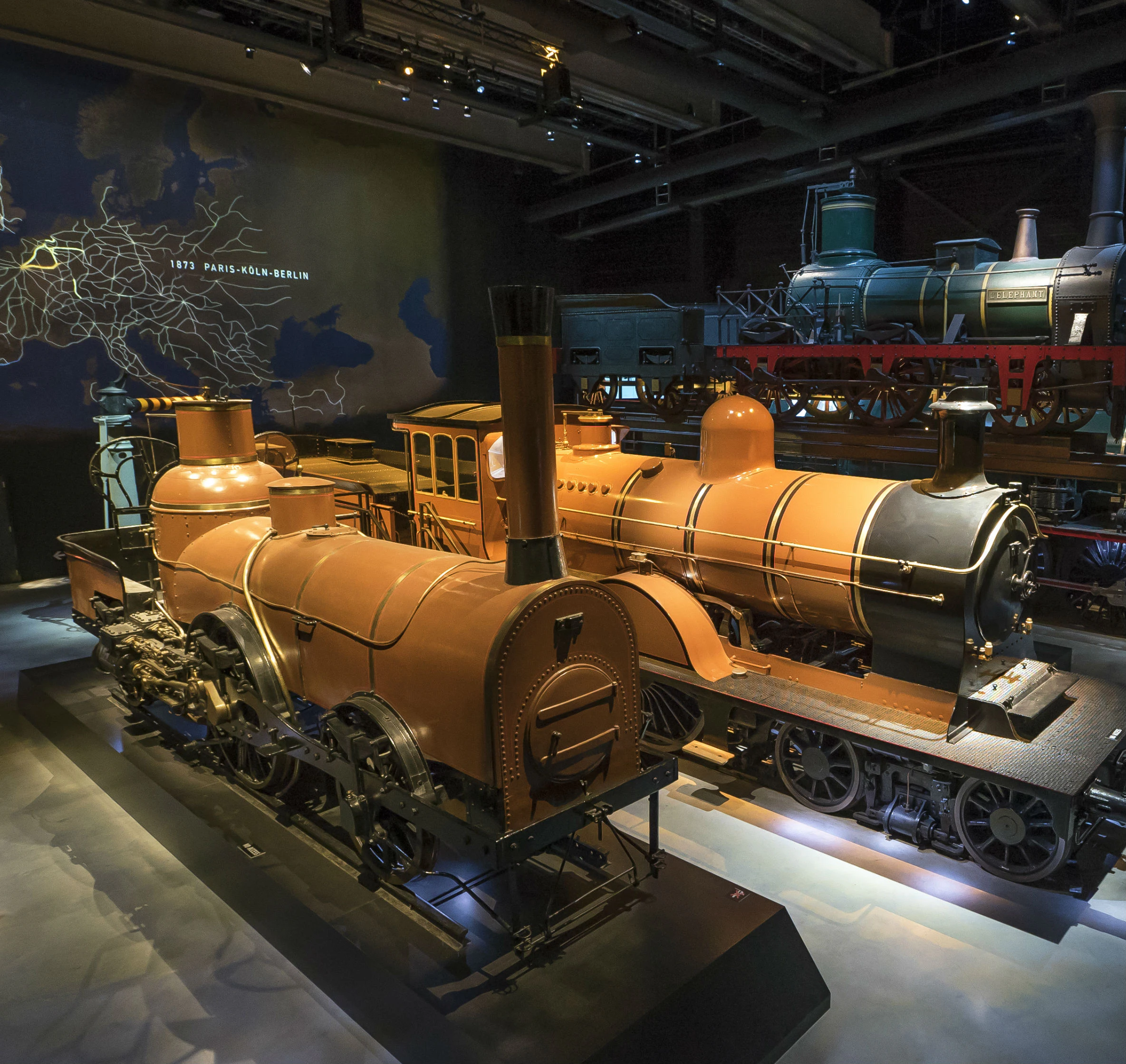 Two antique steam locomotives displayed in a dimly lit museum, with an old railway map on the wall in the background.