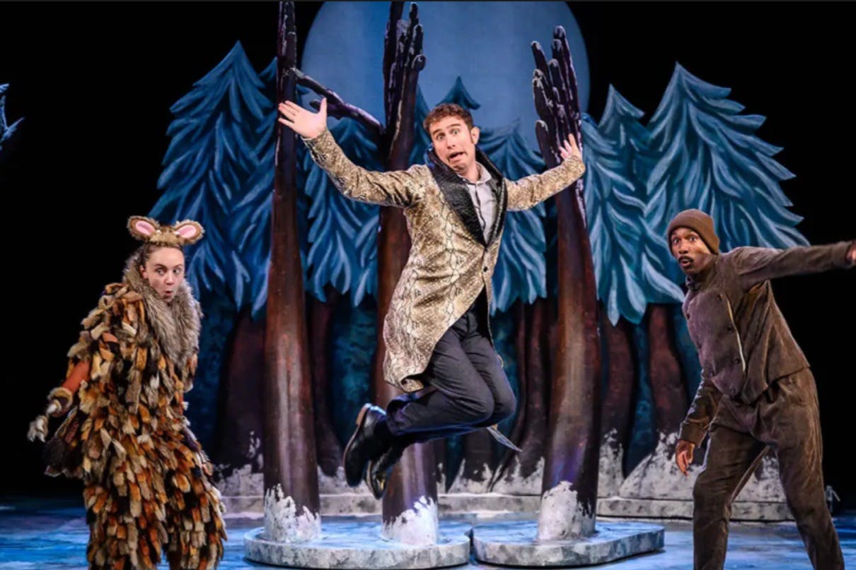 Tři herci v kostýmech vystupují na jevišti se zasněženou lesní kulisou. Jeden herec skáče do vzduchu, zatímco ostatní reagují dramaticky.