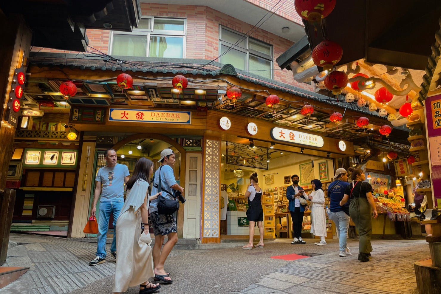People walking in front of a traditional-style shop adorned with red lanterns. The shop sign reads "DAEBAK."