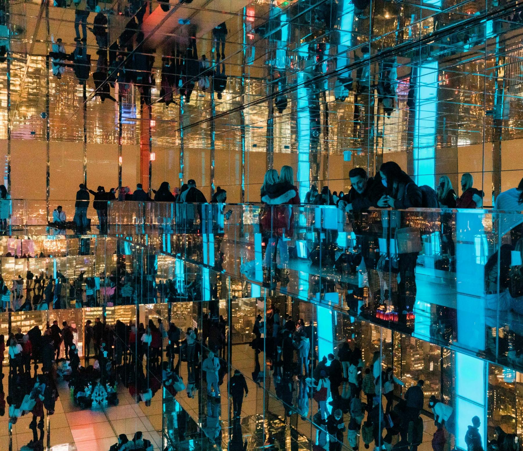 Une pièce dont les murs et le plafond sont recouverts de miroirs reflétant des personnes et des lumières bleues, créant un effet visuel complexe.