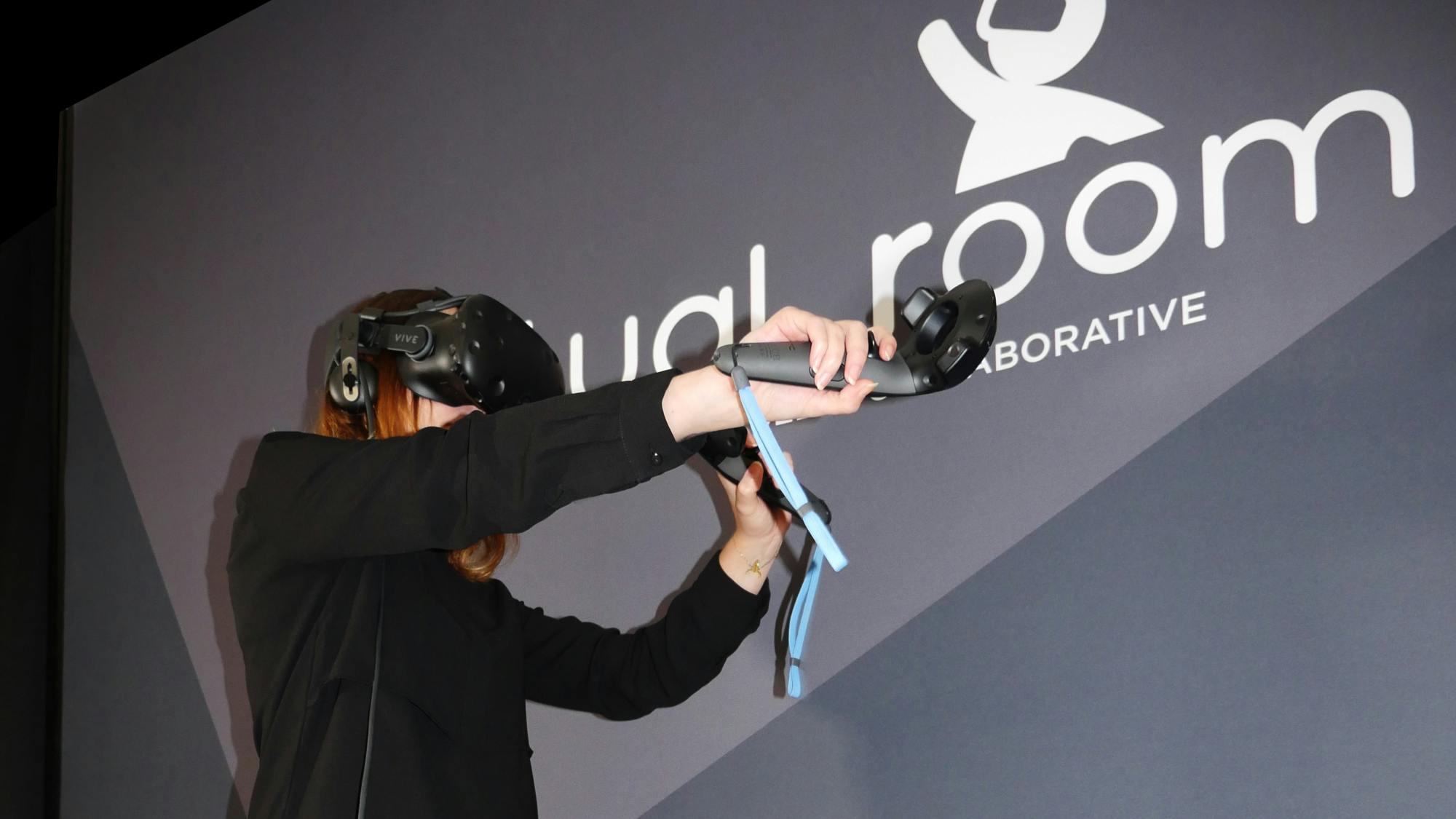 Persoon met een virtual reality-headset en handcontrollers tegen een muur met het logo "Dual Room Laboratories".