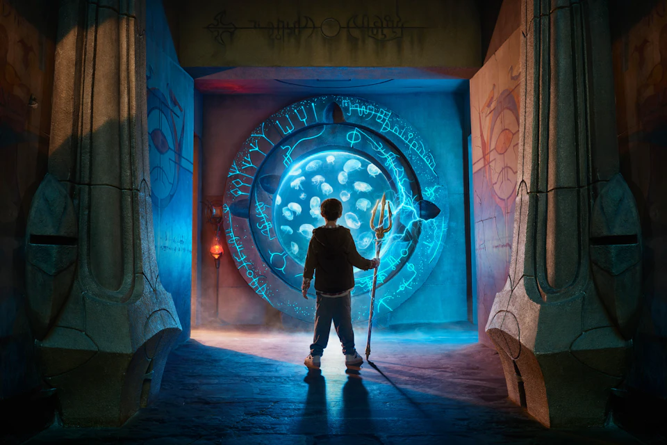 A person holding a trident stands in front of a glowing circular portal with symbols, surrounded by an ancient stone setting.