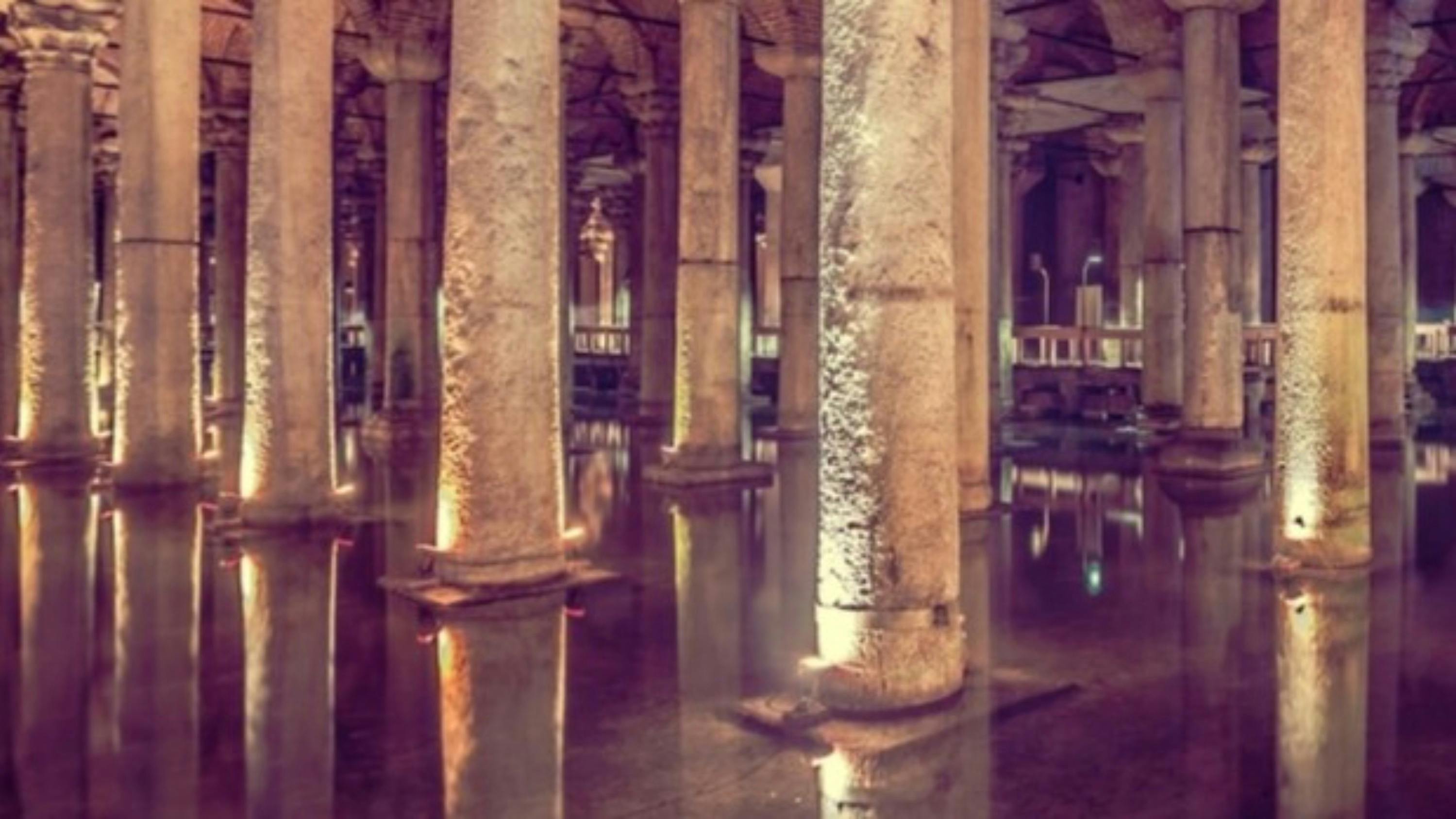 An ancient underground structure with stone columns, reflective water, and warm lighting.