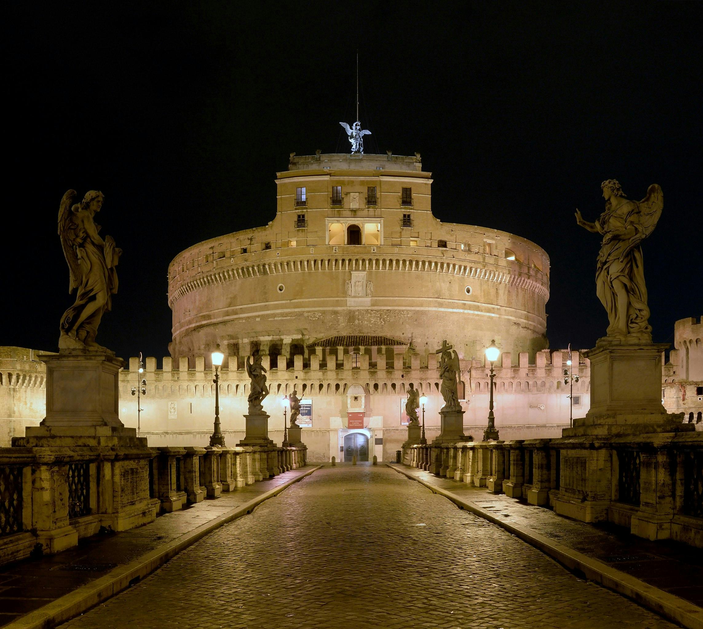 Night view of Castel Sant'Angelo in Rome, featuring illuminated statues, stone pathway, and lampposts leading to the entrance.