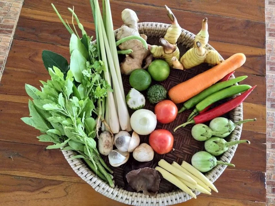 Wicker basket filled with fresh vegetables including lemongrass, carrots, tomatoes, limes, and various greens on a wooden surface.