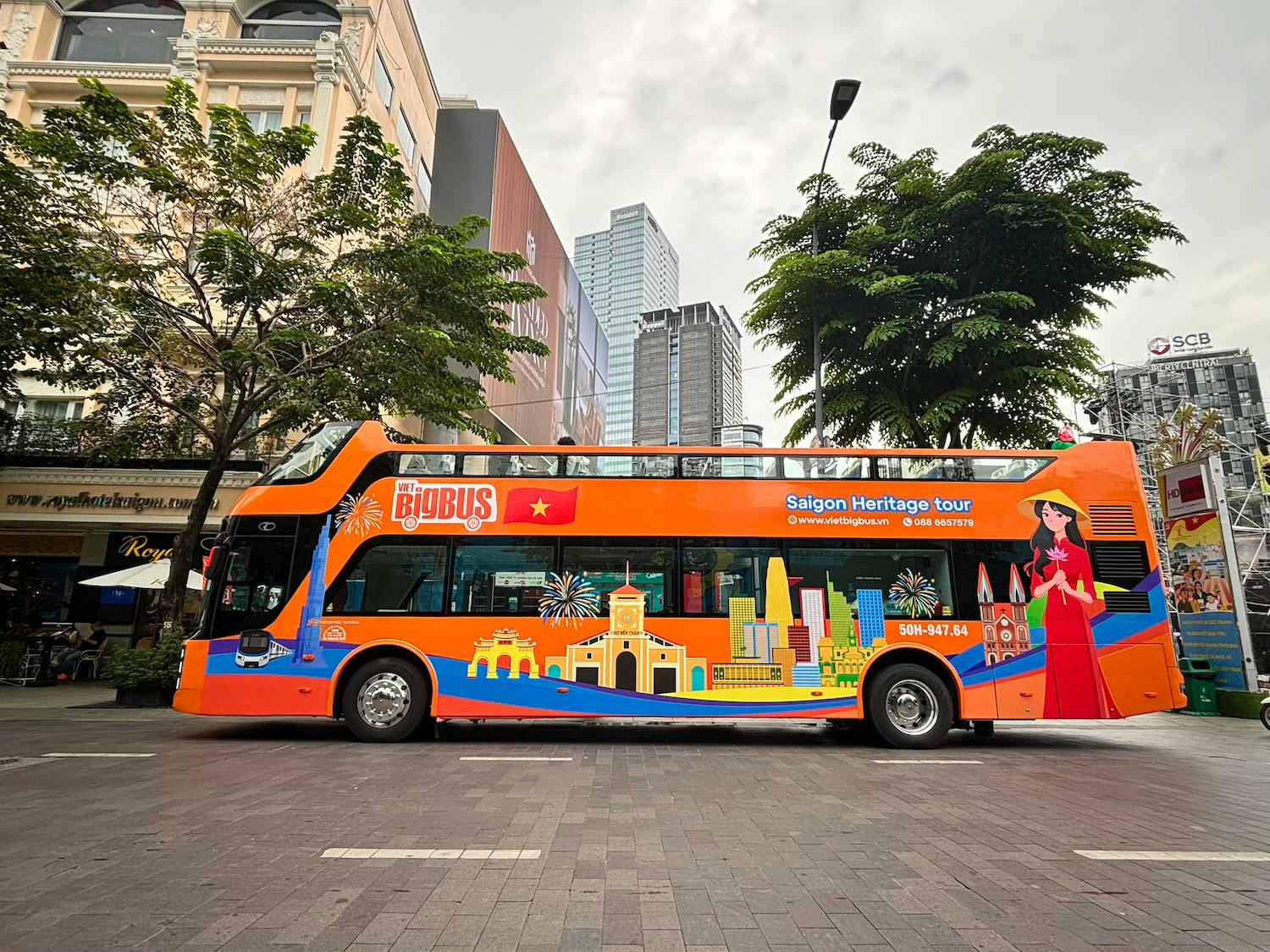 A bright orange, double-decker tour bus with illustrations of Saigon landmarks parked on a street with trees and buildings.