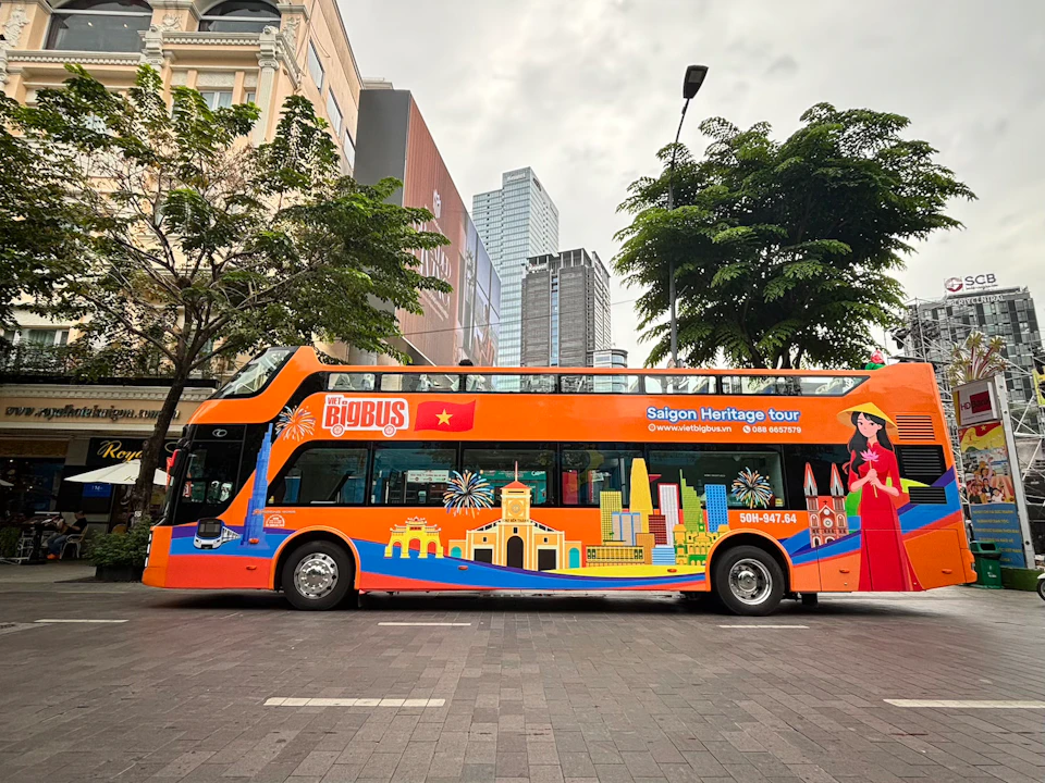 An orange double-decker bus parked on a city street with Saigon Heritage Tour branding and colorful illustrations.