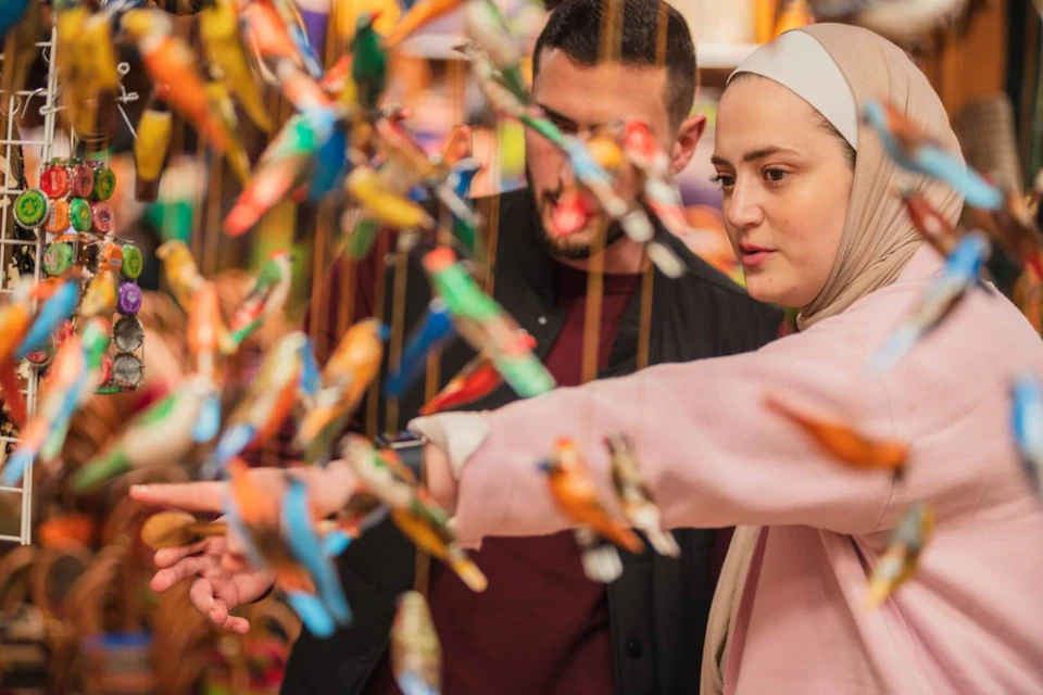Two people, one in a headscarf, are looking at colorful bird ornaments, with one person pointing at them.