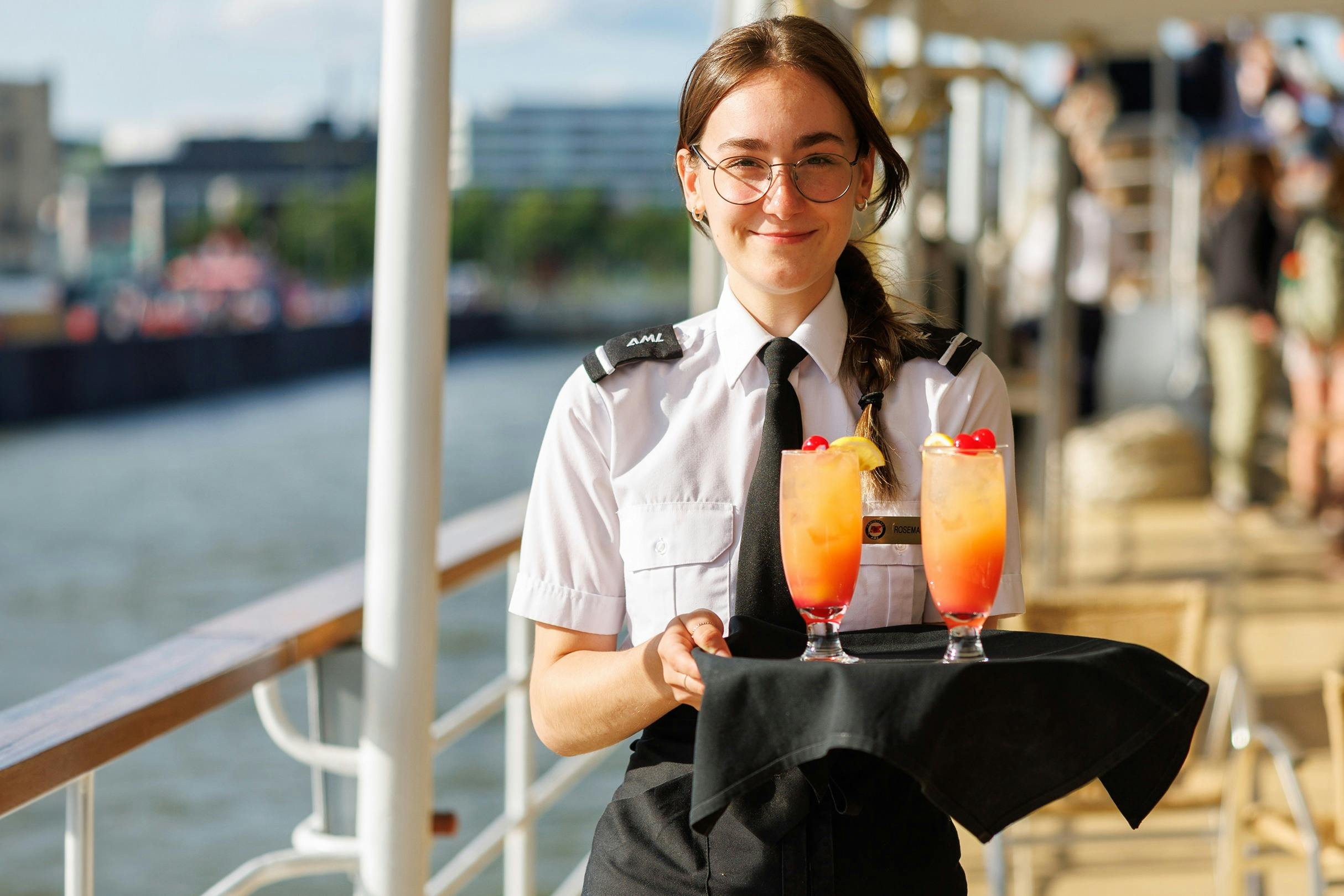 A person in a uniform holding a tray with two vibrant cocktails, standing on a ship's deck with a river and buildings visible.