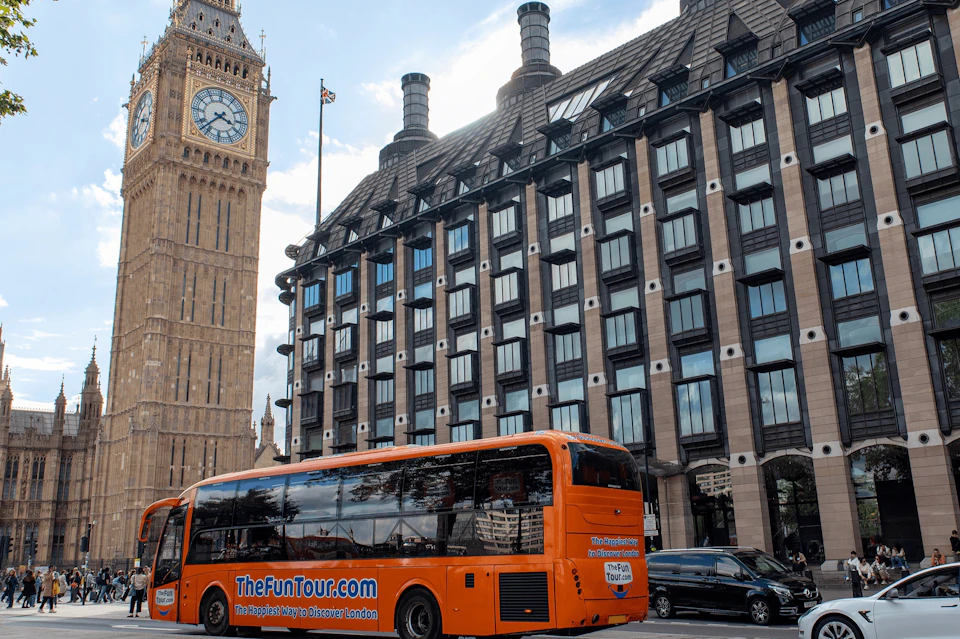 An orange tour bus parked in front of a building with many windows, with Big Ben visible in the background.