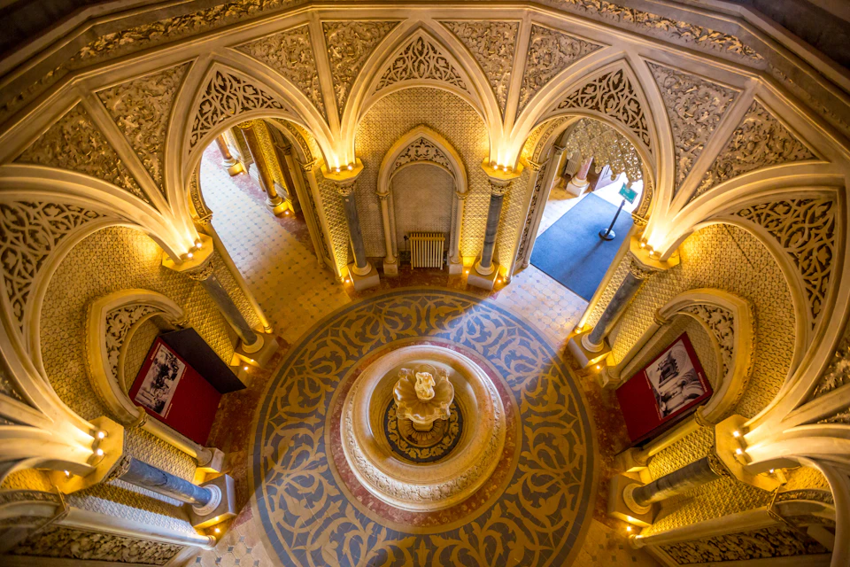 Elaborately decorated hall with Gothic arches, ornate carvings, a central fountain, and golden lighting, viewed from above.