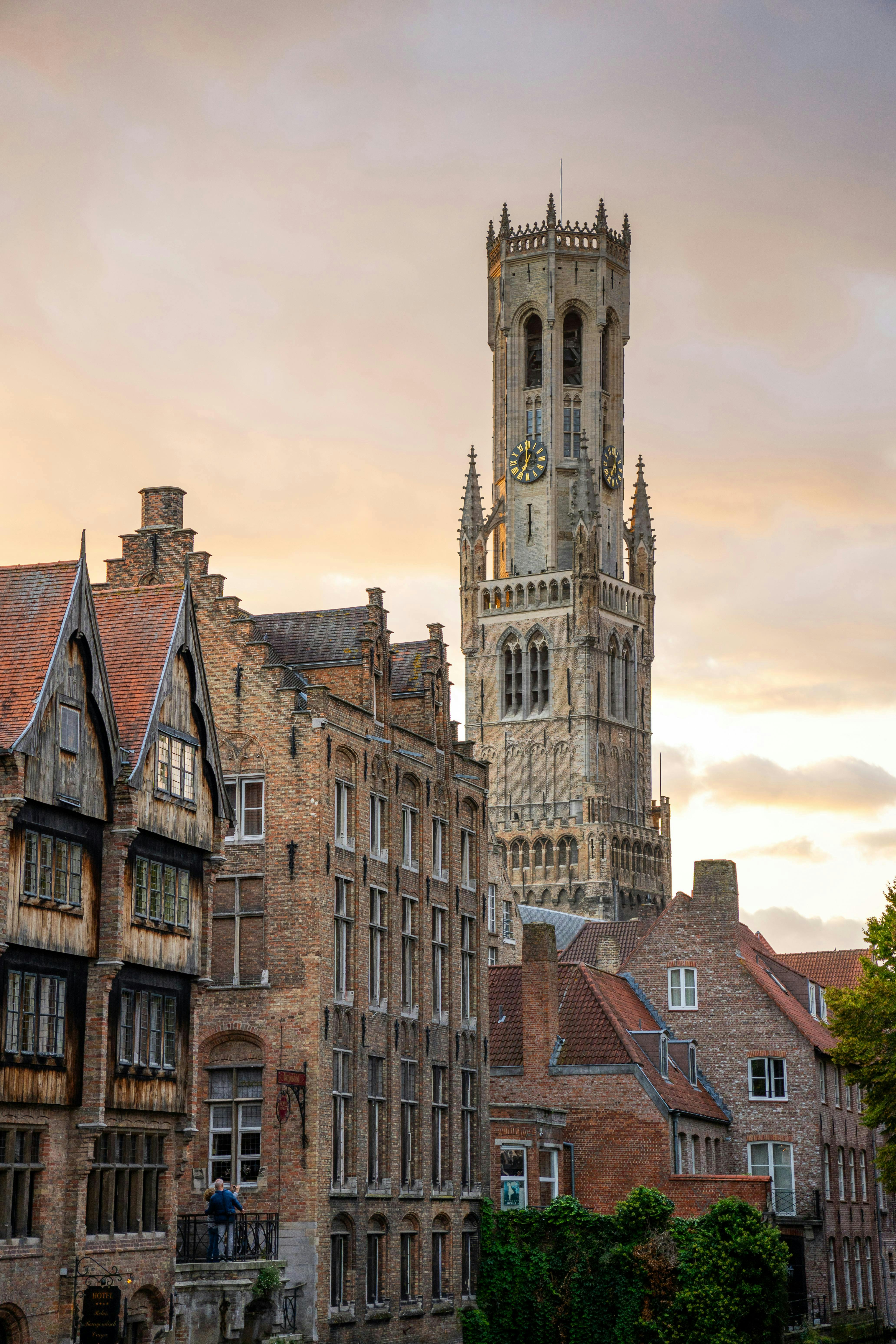 A view of a tall medieval clock tower surrounded by traditional brick buildings under a cloudy sky at sunset.