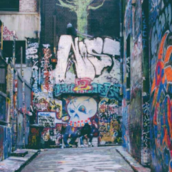 An alleyway covered in colorful graffiti, including a large stylized face and various tags. Some people are present in the background.