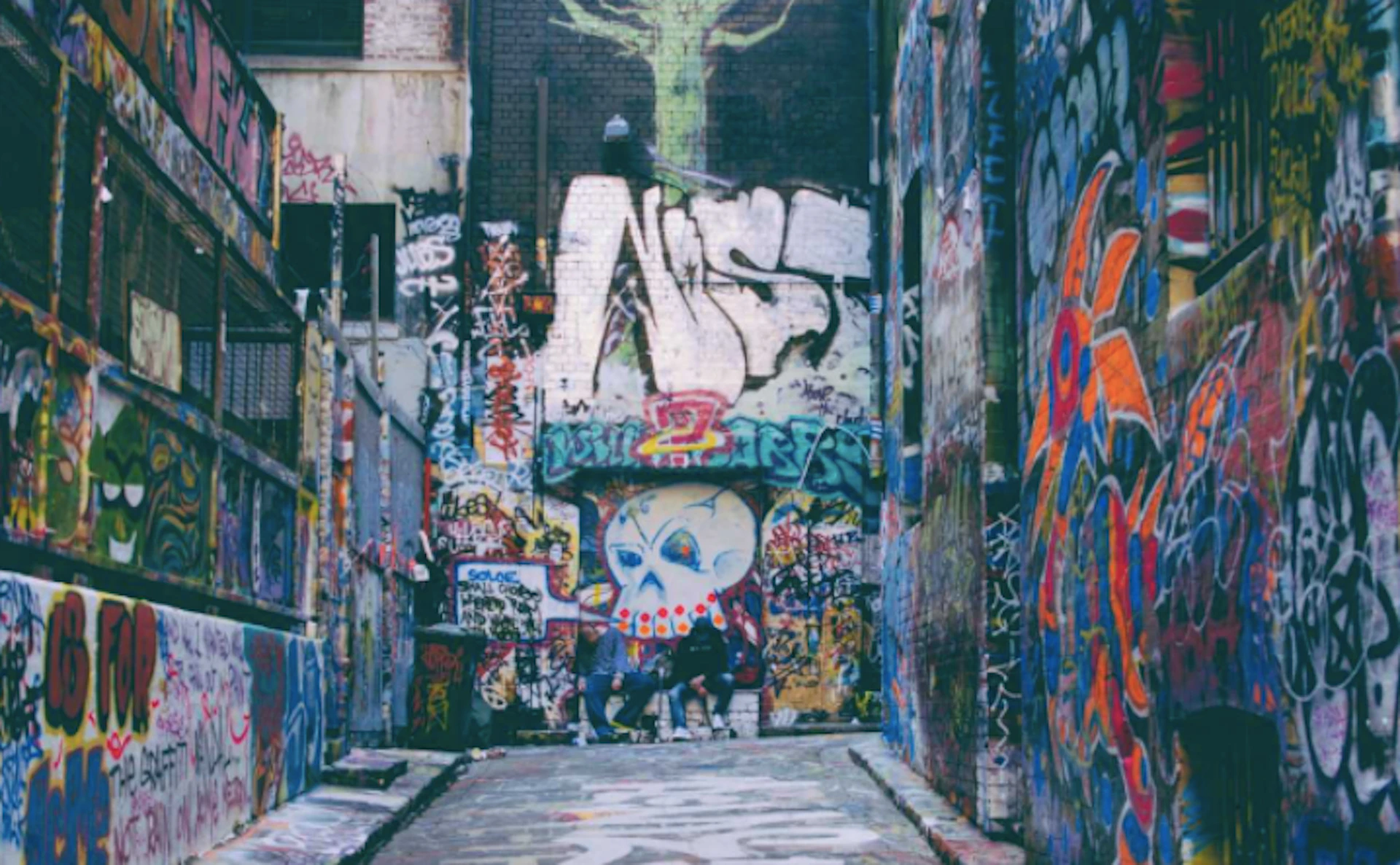 An alleyway covered in colorful graffiti, including a large stylized face and various tags. Some people are present in the background.