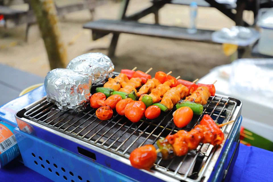 Vegetable and meat skewers, along with foil-wrapped food, grilling on an outdoor barbecue with a picnic bench in the background.