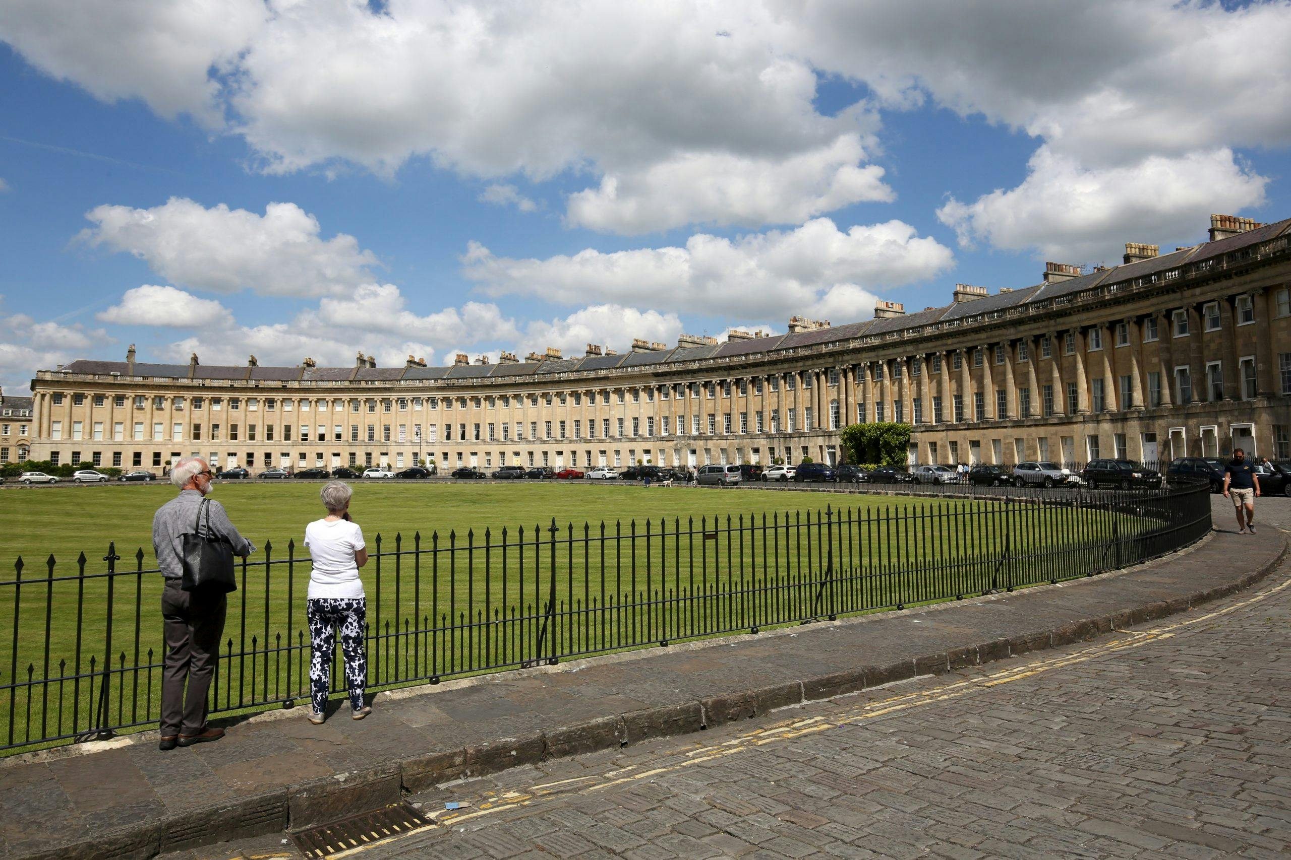 Two people stand in front of a black iron fence, facing a large, curved row of beige terraced houses under a partly cloudy sky.