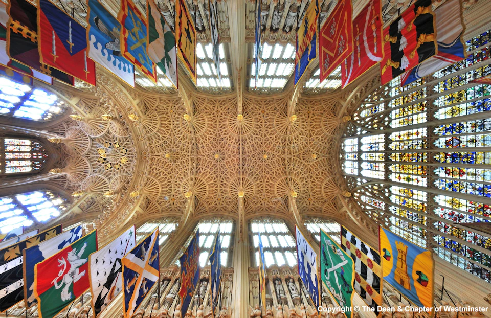 Ornate ceiling of a cathedral adorned with intricate carvings and colorful flags, under large stained glass windows.
