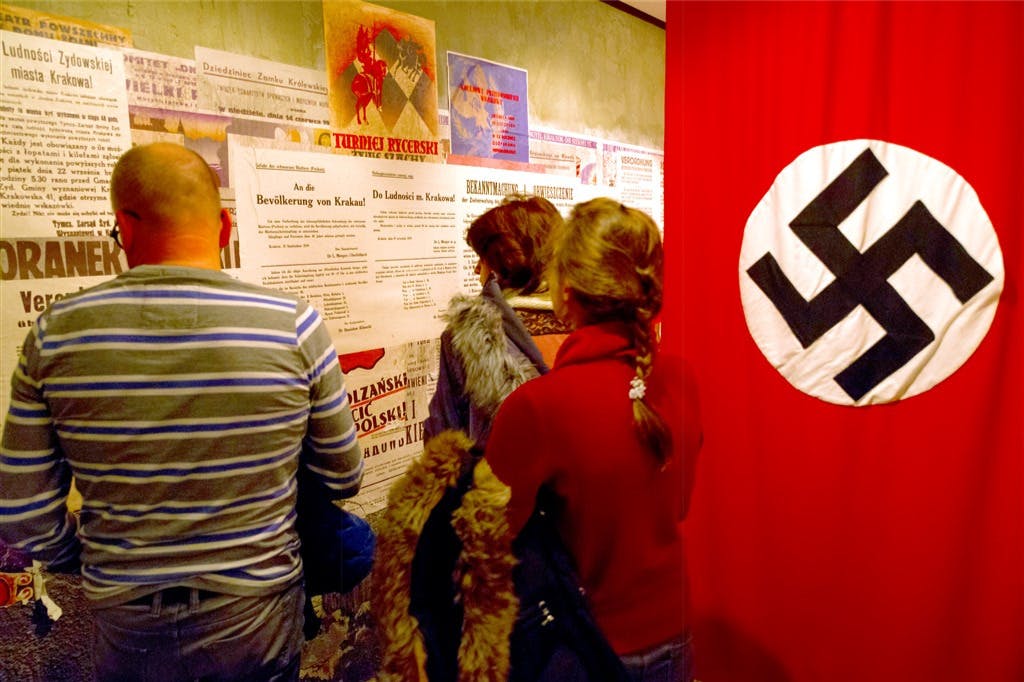人们在墙上阅读历史海报，旁边是一面大红旗，白色圆圈上有一个黑色的纳粹标志。