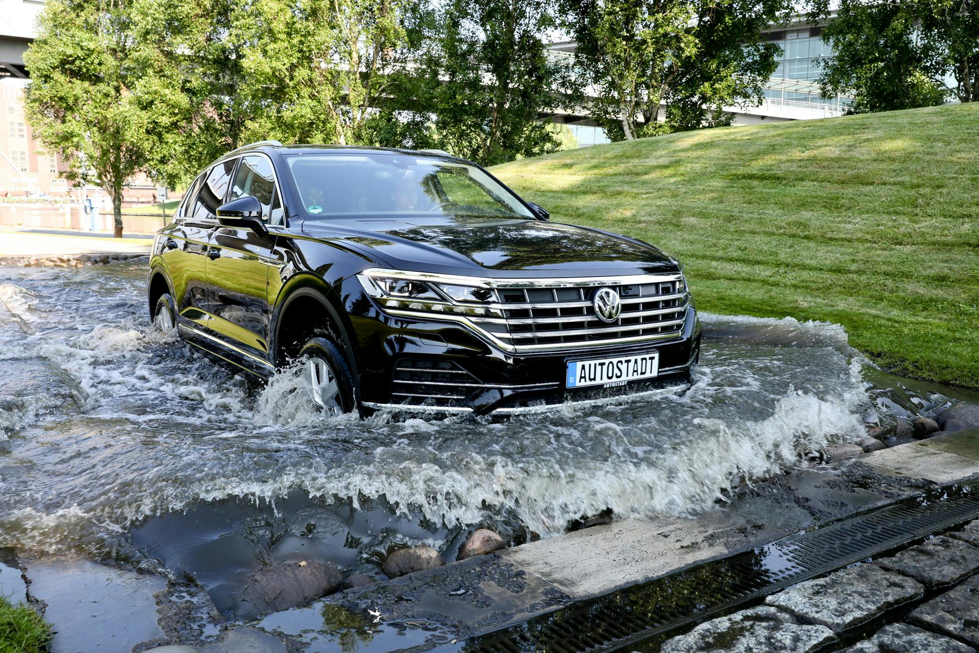 A black Volkswagen SUV driving through a shallow water course with trees and a grassy slope in the background.